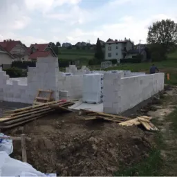 Budowa domu jednorodzinnego w trakcie wznoszenia ścian z bloczków betonowych, widoczne materiały budowlane i narzędzia na placu budowy.