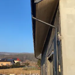 Fragment ściany budynku w trakcie ocieplania, widoczne płyty izolacyjne, rynna, podbitka drewniana, w tle zabudowa jednorodzinna i las.