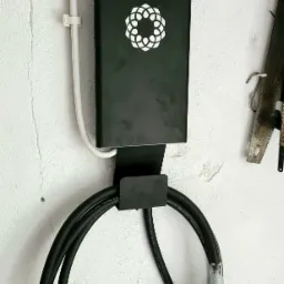 Czarna stacja ładowania pojazdów elektrycznych z białym logo, zamontowana na białej, teksturowanej ścianie garażu, z widocznym kablem w ochronnej folii i narzędziami w tle.