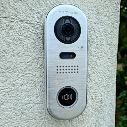 Nowoczesny, srebrny panel wideodomofonu Vidos zamontowany na szarej, chropowatej ścianie zewnętrznej budynku, z widoczną kamerą, przyciskiem dzwonka i symbolem kłódki.