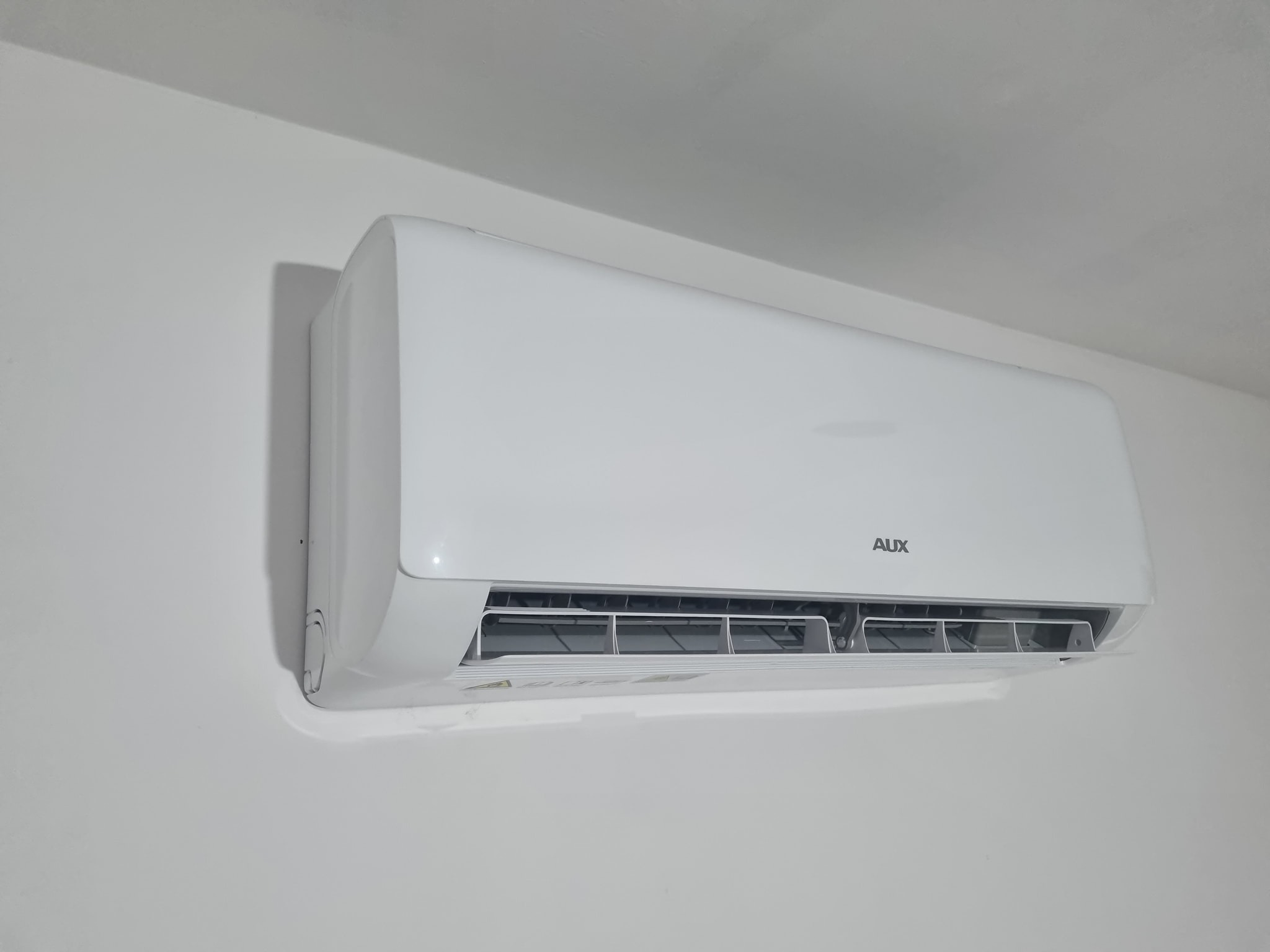 Biała jednostka klimatyzacji marki AUX zamontowana na białej ścianie w Lubinie, widok z boku i lekko od dołu.