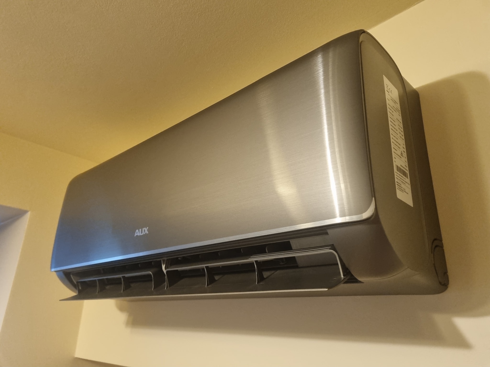 Szary klimatyzator AUX zamontowany na kremowej ścianie, widoczne żaluzje wylotu powietrza i etykieta z parametrami technicznymi po prawej stronie.