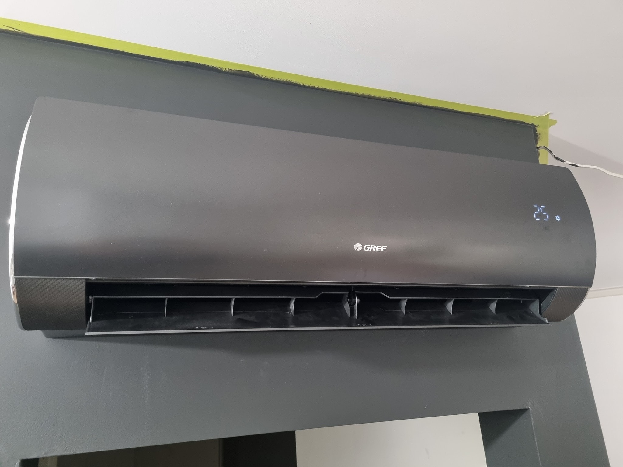 Czarna jednostka wewnętrzna klimatyzatora GREE zamontowana na szarej ścianie, widoczna taśma malarska na styku ściany i sufitu.