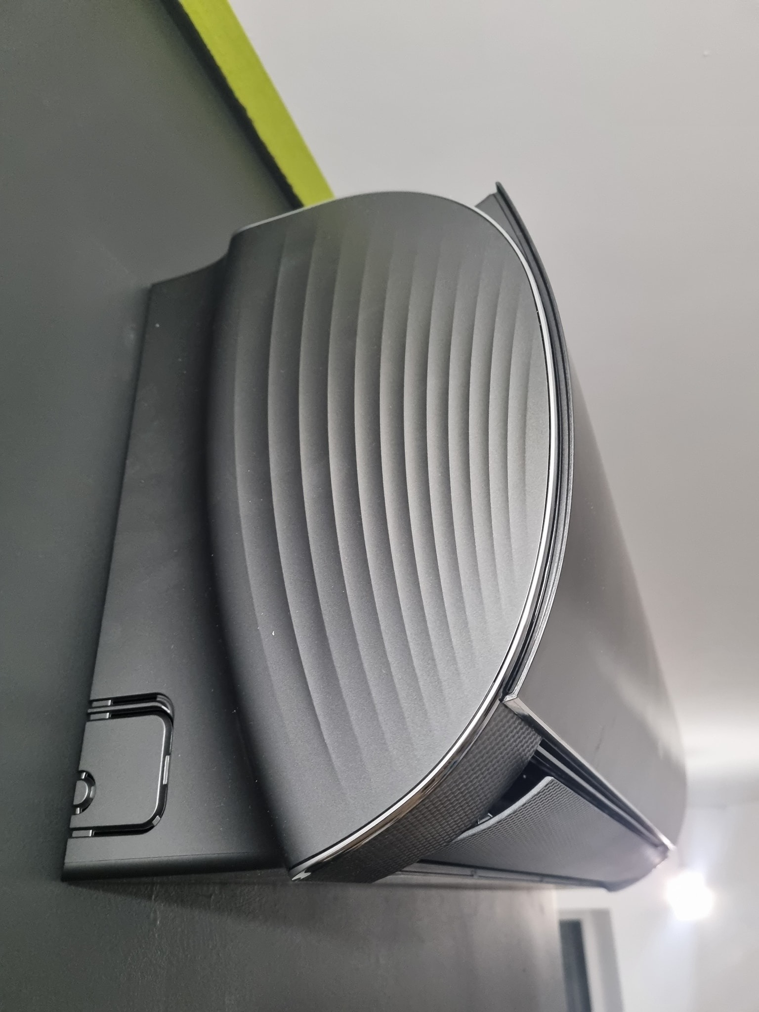 Czarny klimatyzator ścienny z falistą strukturą obudowy, zamontowany na ciemnej ścianie z zielonym akcentem przy suficie, widok z dołu pod kątem.