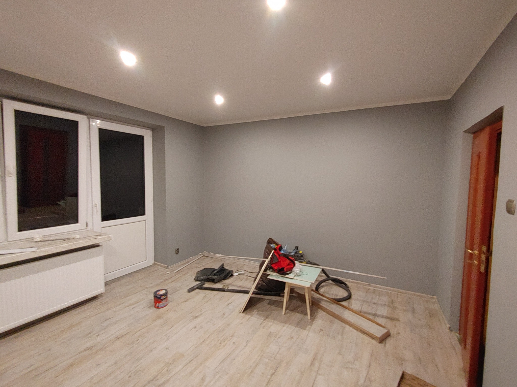 Pokój w trakcie prac: szare ściany, drewnopodobna podłoga, białe okno balkonowe i drzwi, grzejnik, rozstawione narzędzia i materiały budowlane, widoczne ślady po malowaniu.