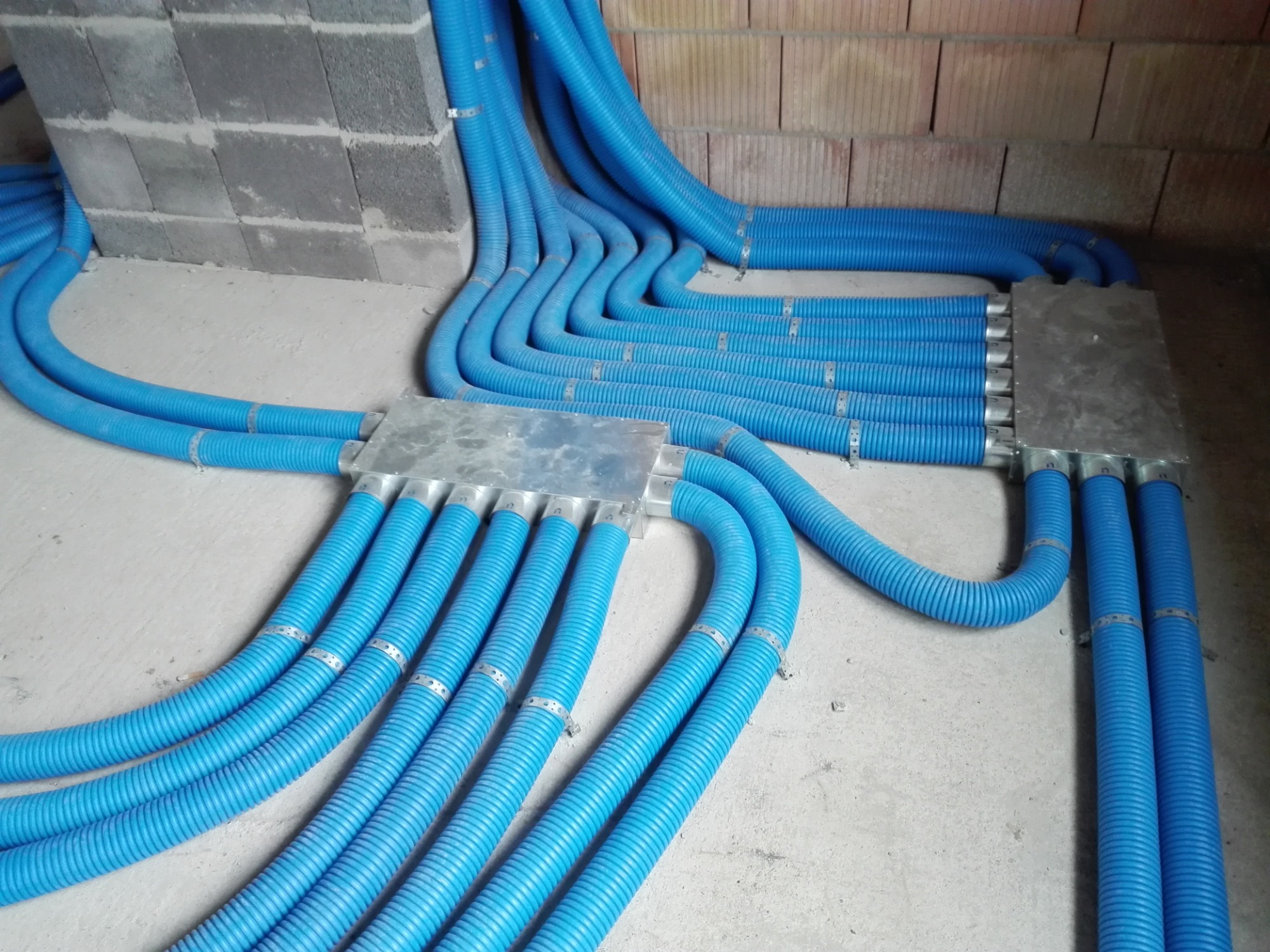 Niebieskie, karbowane rury prowadzone wzdłuż betonowej podłogi i ściany z pustaków, połączone metalowymi skrzynkami rozdzielczymi.