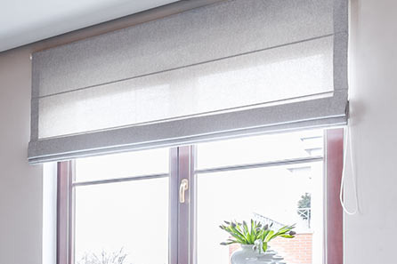 Szara tekstylna osłona okienna w stylu rzymskim, częściowo opuszczona, z widocznym sznurkiem sterującym, na tle okna z bukietem tulipanów w wazonie.