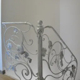 Biała balustrada schodowa z metalu, zdobiona motywem liści winogron, z drewnianą podłogą w tle, ujęcie z dołu.