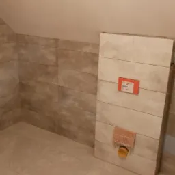 Ściana w łazience wyłożona płytkami imitującymi drewno, z widoczną instalacją sanitarną i fragmentem podłogi wyłożonej jasnymi płytkami.