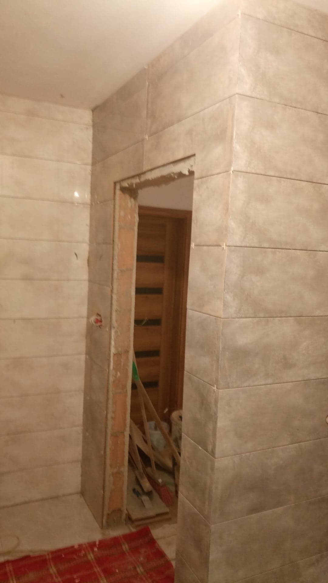 Ściana łazienki wyłożona poziomymi płytkami w odcieniach szarości, widoczne niedokończone obramowanie drzwi z cegły oraz fragment podłogi z czerwoną szmatą. W tle widoczne narzędzia i drzwi.