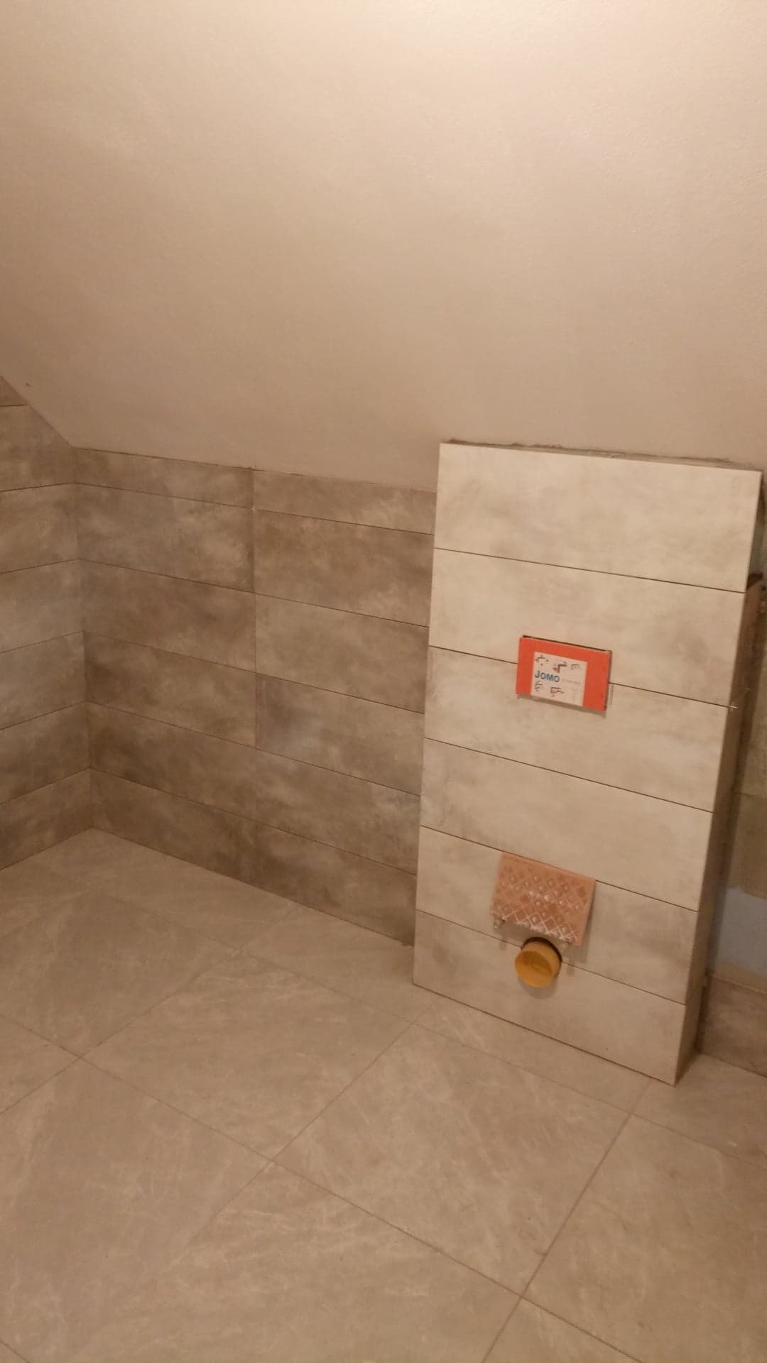 Ściana w łazience wyłożona płytkami imitującymi drewno, z widoczną instalacją sanitarną i fragmentem podłogi wyłożonej jasnymi płytkami.