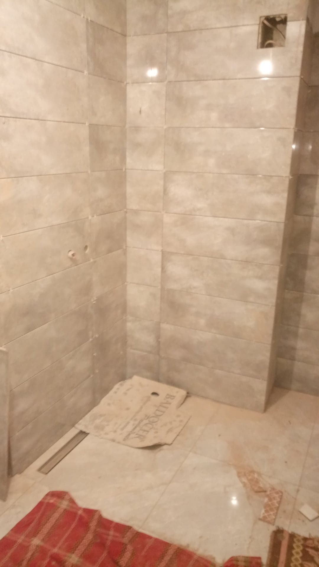 Ściany łazienki wyłożone beżowymi, prostokątnymi płytkami, widoczne otwory na instalacje, fragment kratki odpływowej w podłodze, resztki zaprawy i zabrudzenia po remoncie, oraz kartonowa podkładka...