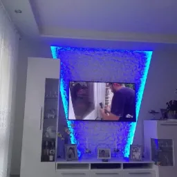 Nowoczesna ściana telewizyjna z podświetleniem LED w kolorze niebieskim, białe meble RTV z szufladami i półkami, dekoracyjne ramki ze zdjęciami.