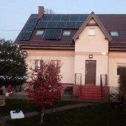 Rodzinny dom jednorodzinny z brązowym dachem i panelami słonecznymi, oświetlony choinką na trawniku, sfotografowany o zmierzchu.