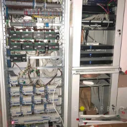 Dwie szafy elektryczne z widocznymi kablami, bezpiecznikami i sterownikami, jedna z elementami sieciowymi Lanberg, w tle widoczne ślady po pracach budowlanych.