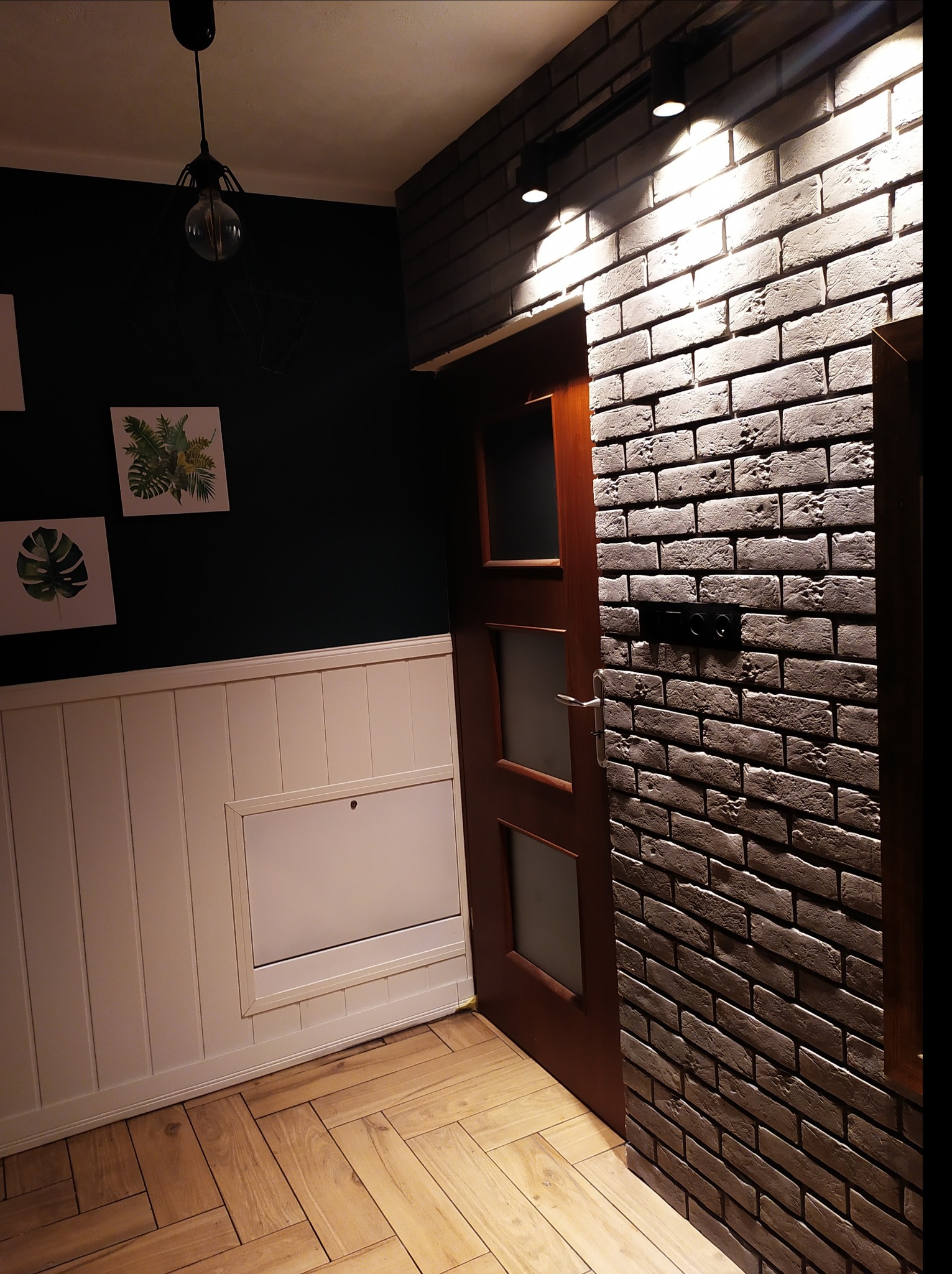 Wnętrze z szarą ścianą z cegły dekoracyjnej, drewnianą stolarką drzwiową z przeszkleniami, parkietem ułożonym we wzór jodełki oraz dolną częścią ściany wykończoną białą boazerią panelową.