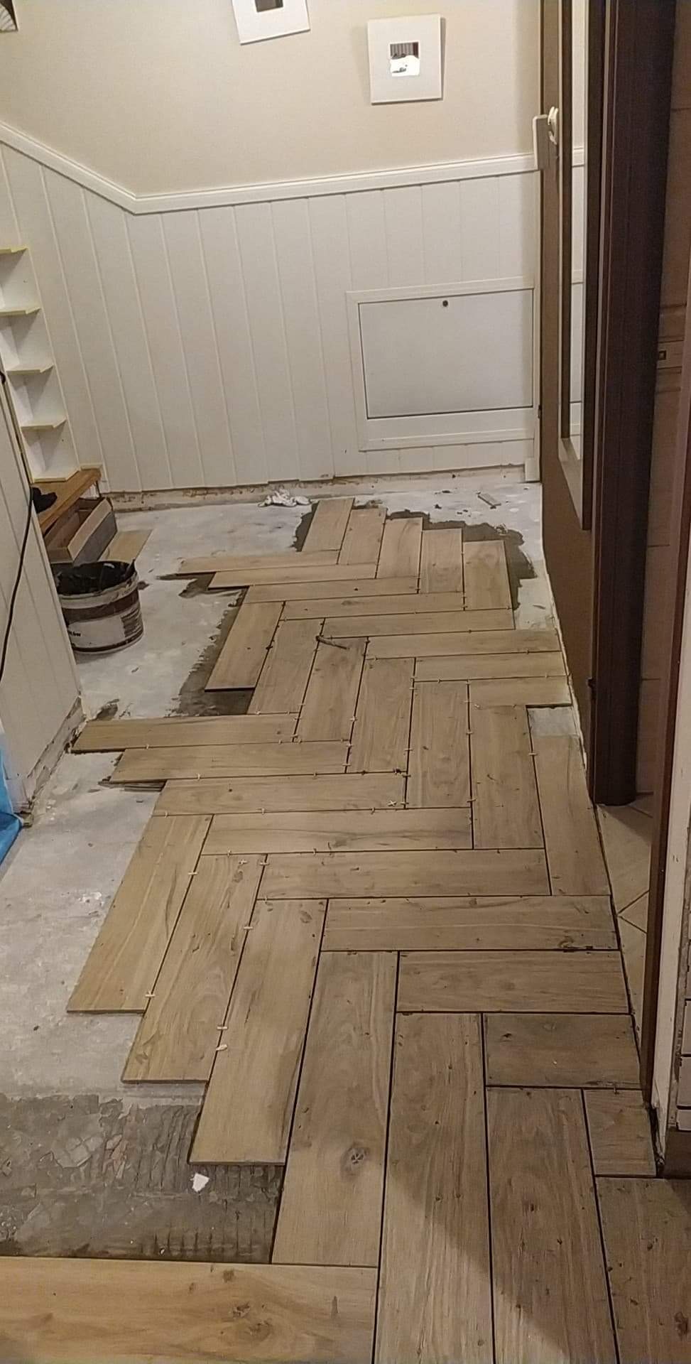 Układanie drewnopodobnych płytek w jodełkę na podłodze, widoczne krzyżyki dystansowe, ślady kleju i wiadro z zaprawą w tle.