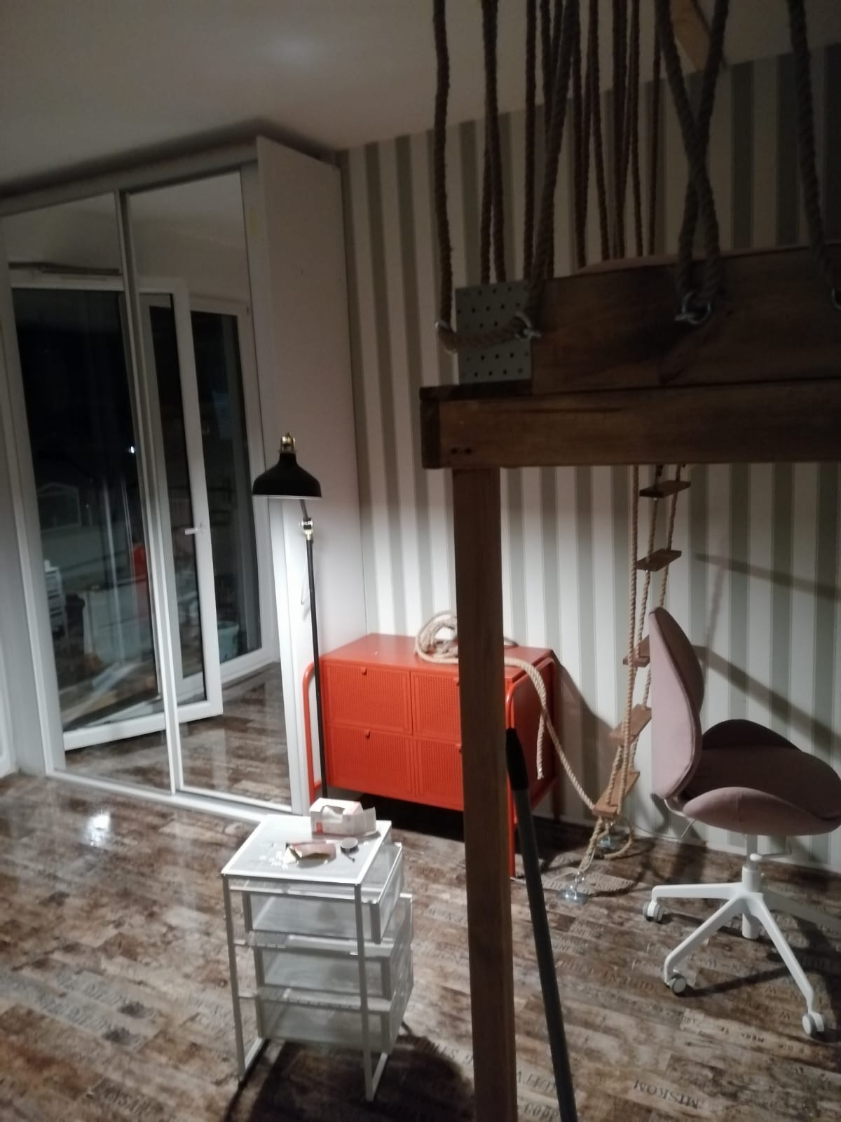 Pokój z drewnianym łóżkiem na antresoli z linami, pomarańczową komodą, biurkiem na kółkach, krzesłem biurowym i przesuwnymi drzwiami balkonowymi.