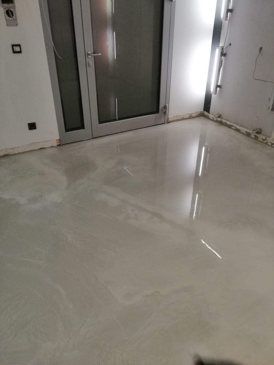 Wylany, samopoziomujący podkład podłogowy w pomieszczeniu z aluminiowymi drzwiami i listwami przypodłogowymi w trakcie montażu.