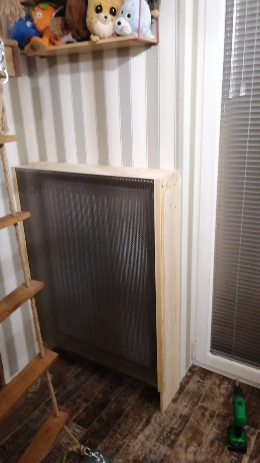 Drewniana obudowa grzejnika z metalową siatką w pokoju dziecięcym, widoczna drabinka sznurkowa i zabawki na półce, obok okno z żaluzjami i wkrętarka na podłodze.