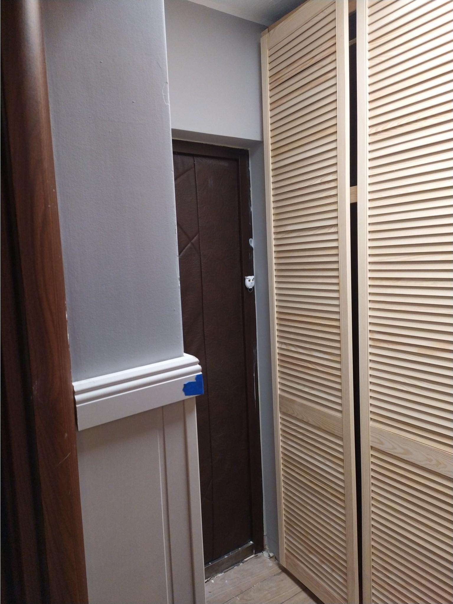 Szare wnętrze z drewnianymi, lamelowymi drzwiami przesuwnymi, brązowymi drzwiami wejściowymi i listwą przypodłogową zabezpieczoną niebieską taśmą malarską.