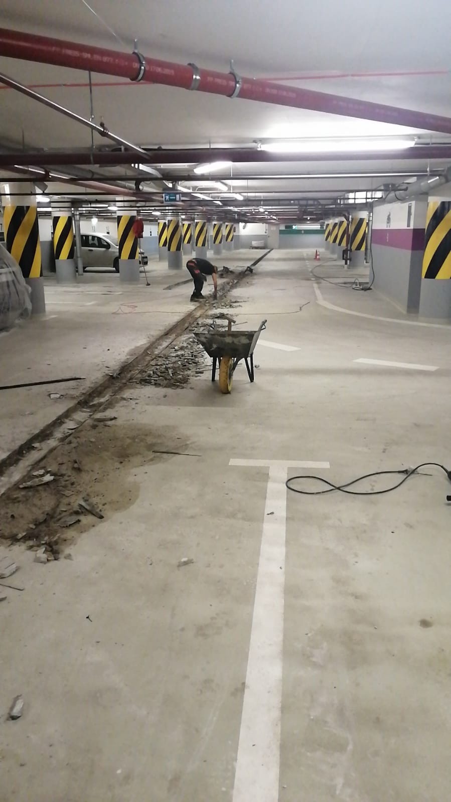 Pracownik w garażu podziemnym skuwa betonową posadzkę wzdłuż linii, gruz w taczce, widoczne instalacje pod sufitem.