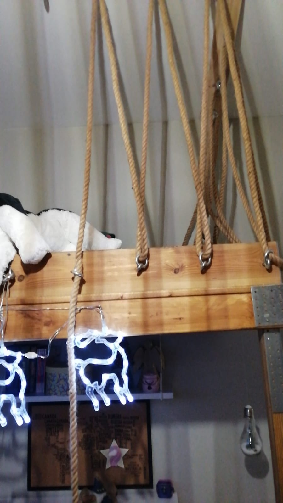 Drewniana konstrukcja łóżka na linach z oświetleniem dekoracyjnym w kształcie reniferów, widok z dołu na półkę z książkami i dekoracjami.