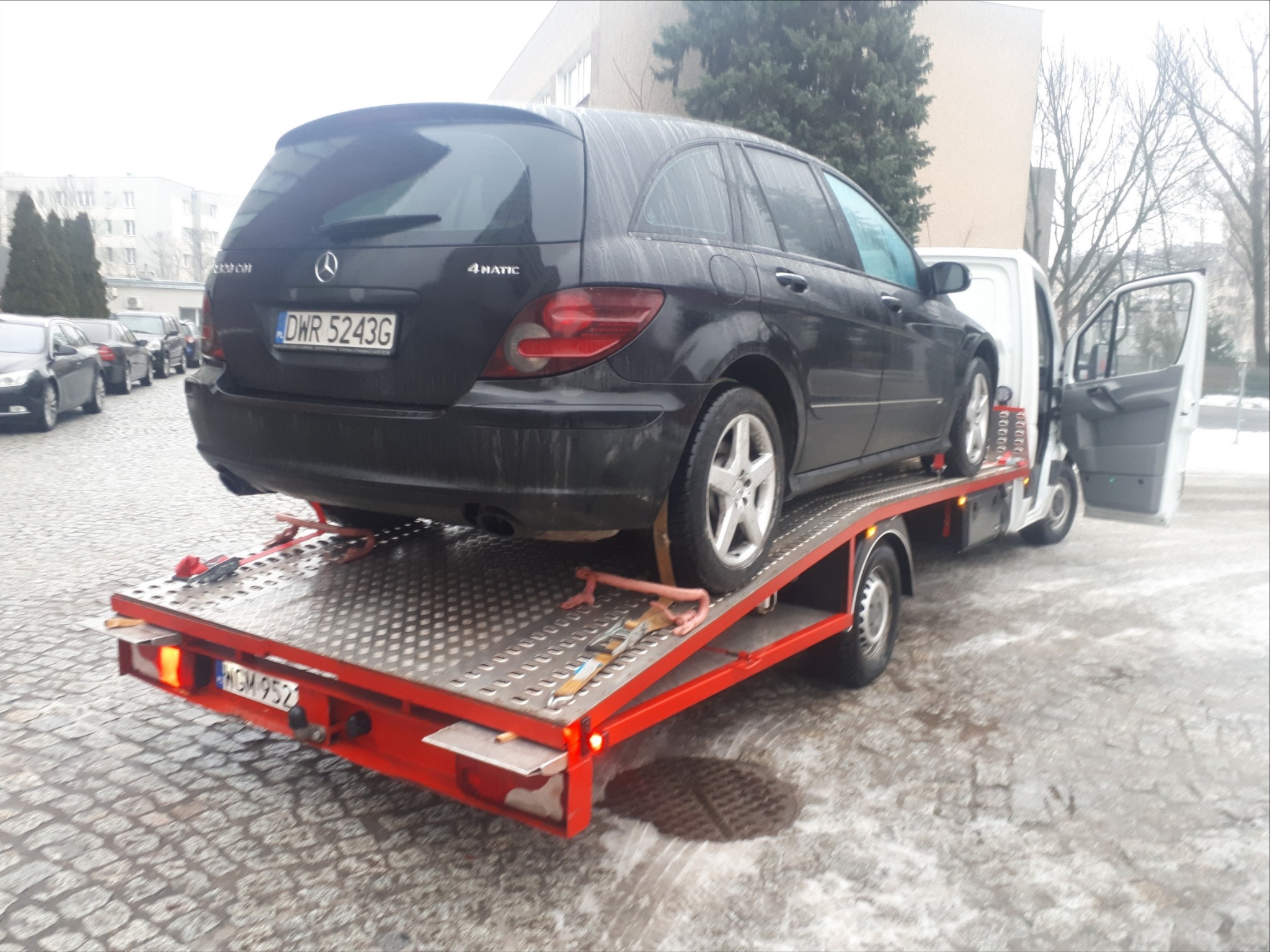 Czarny samochód osobowy Mercedes-Benz 4MATIC na czerwonej platformie transportowej, zabezpieczony pasami, widoczny numer rejestracyjny DWR 5243G, biały samochód dostawczy z otwartymi drzwiami w tle...