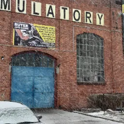 Budynek warsztatu samochodowego z cegły w zimowej scenerii, z niebieską bramą i szyldami reklamowymi, częściowo zasłonięty przez zaparkowany, ośnieżony samochód podczas opadów śniegu.