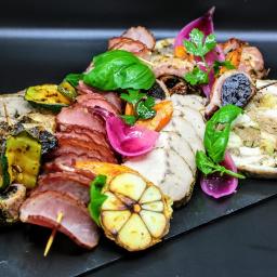 Restauracja Stare Kino - Dekoracyjna kompozycja wędlin, mięs pieczonych, warzyw grillowanych i marynowanych, podana na czarnym kamiennym talerzu.