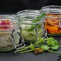 Restauracja Stare Kino - Trzy słoiki z zakrętkami na czarnej kamiennej płycie. Słoik po lewej z białawą pastą i różowymi kiełkami, środkowy z zielonkawą pastą i koperkiem, a po prawej pomarańczowe warzywa w oleju. Obok...