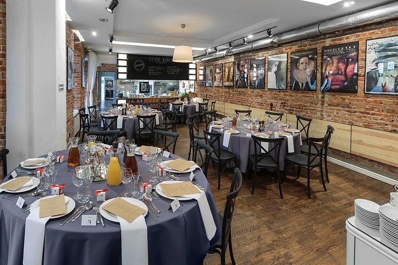 Sala restauracyjna przygotowana na uroczystość, okrągłe stoły z szarymi obrusami, czarne krzesła, nakrycia z białymi serwetkami i upominkami dla gości, na ścianach plakaty filmowe, widoczna tablica...