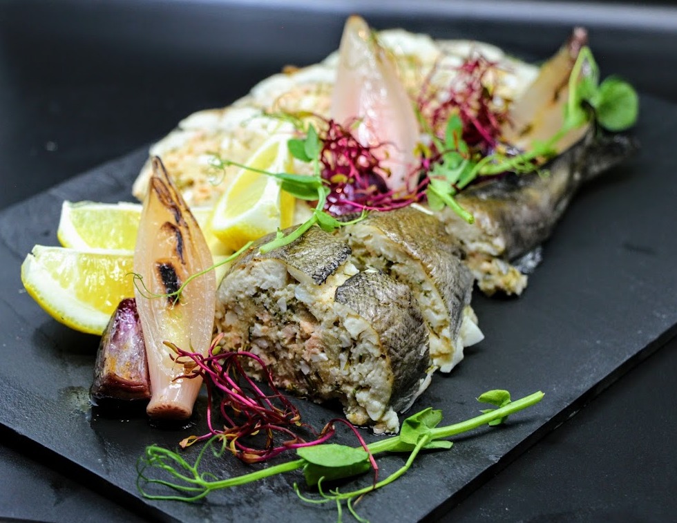 Elegancko podana, faszerowana ryba z cytryną i grillowaną cebulą na czarnym, kamiennym talerzu, udekorowana świeżymi ziołami.