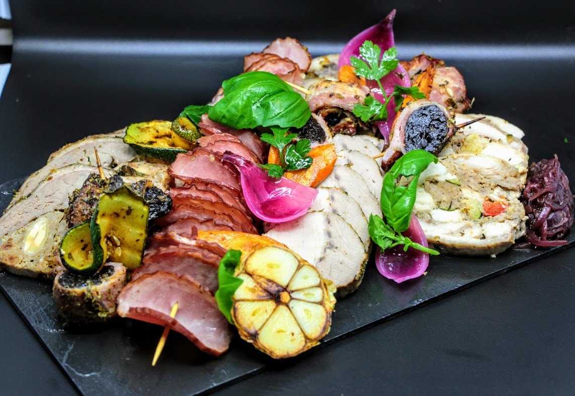 Dekoracyjna kompozycja wędlin, mięs pieczonych, warzyw grillowanych i marynowanych, podana na czarnym kamiennym talerzu.