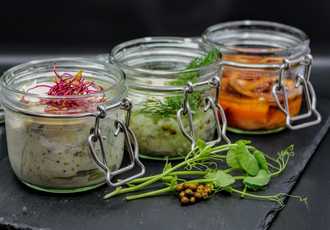 Trzy słoiki z zakrętkami na czarnej kamiennej płycie. Słoik po lewej z białawą pastą i różowymi kiełkami, środkowy z zielonkawą pastą i koperkiem, a po prawej pomarańczowe warzywa w oleju. Obok...