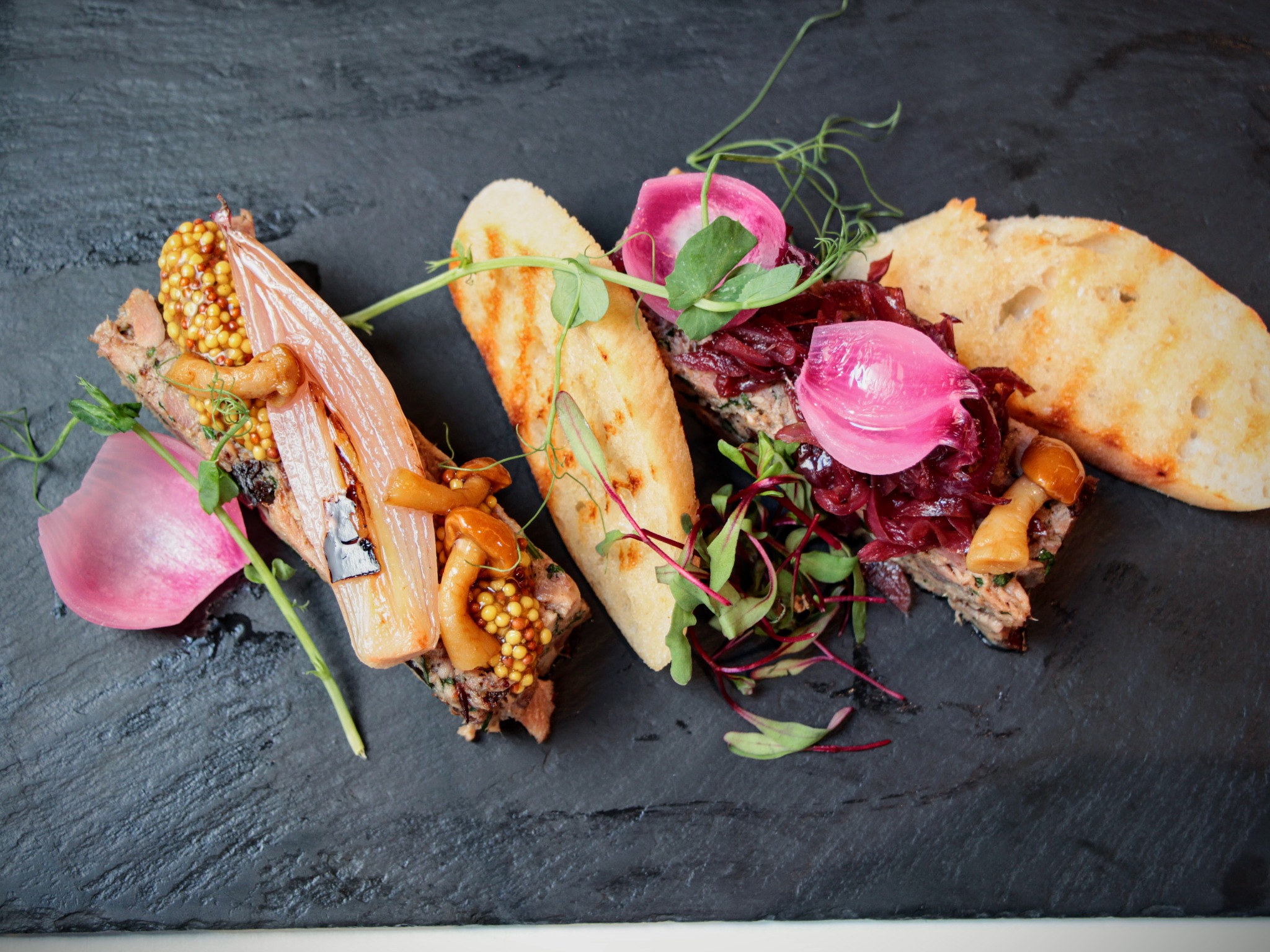 Elegancko podane pasztety z grzankami, musztardą francuską, karmelizowaną cebulą i dekoracjami z mikro ziół na czarnym, kamiennym talerzu.