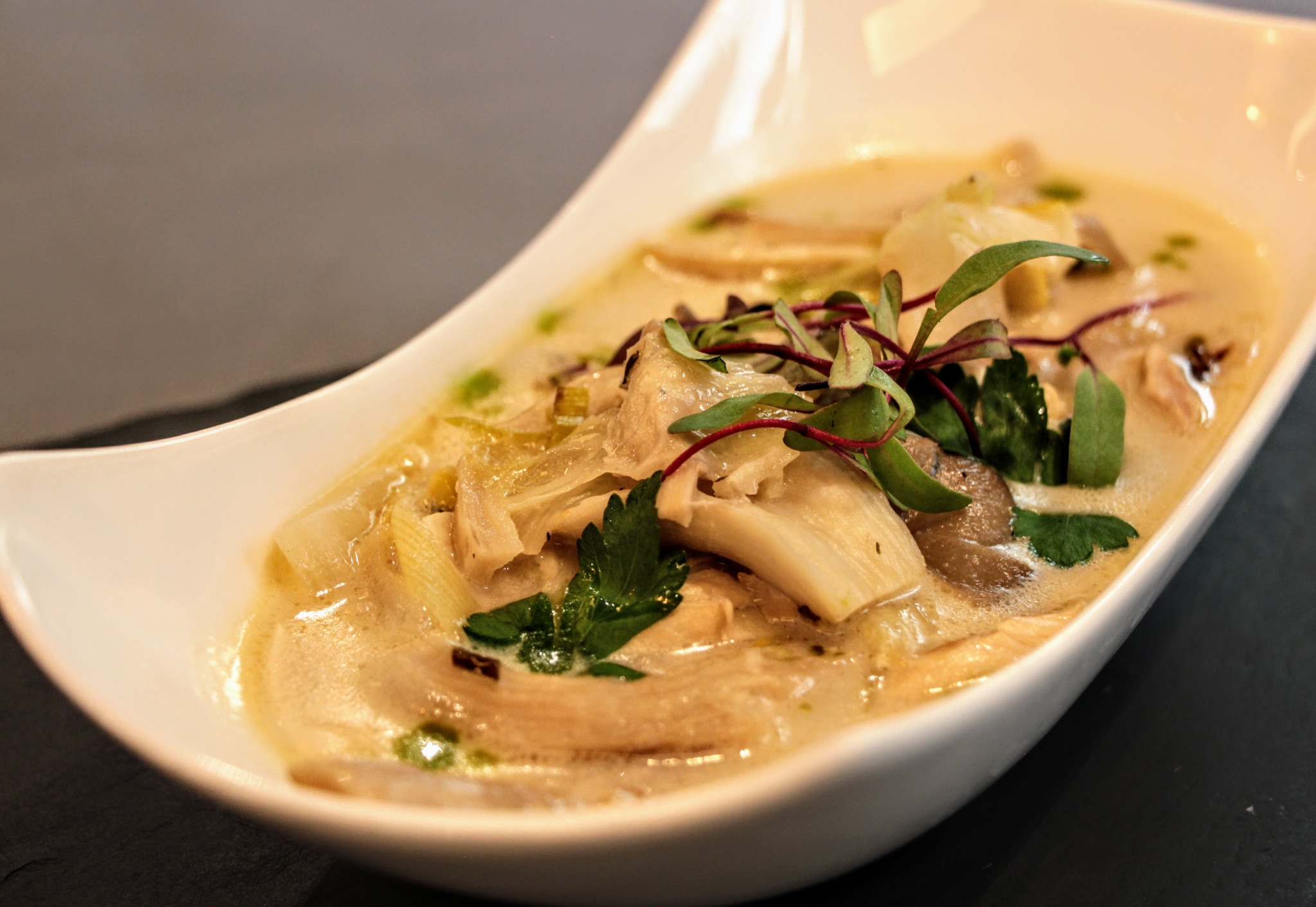 Kremowa zupa z kawałkami grzybów i warzyw, udekorowana świeżymi ziołami, podana w nowoczesnej, białej misce o asymetrycznym kształcie.