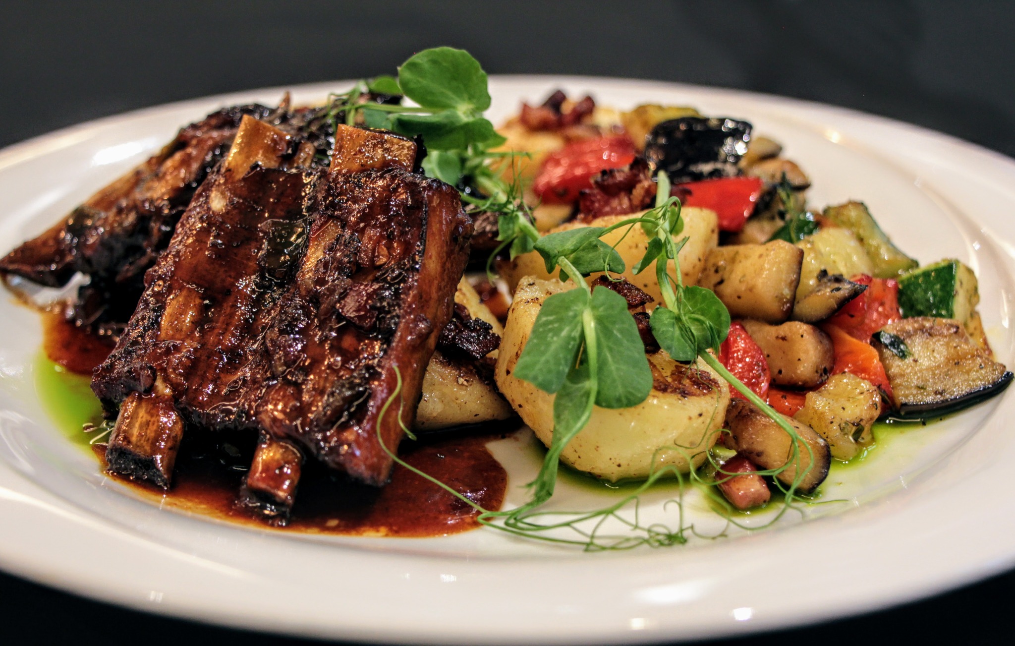 Glazurowane żeberka BBQ podane z pieczonymi ziemniakami, papryką, cukinią i kiełkami groszku na białym talerzu.