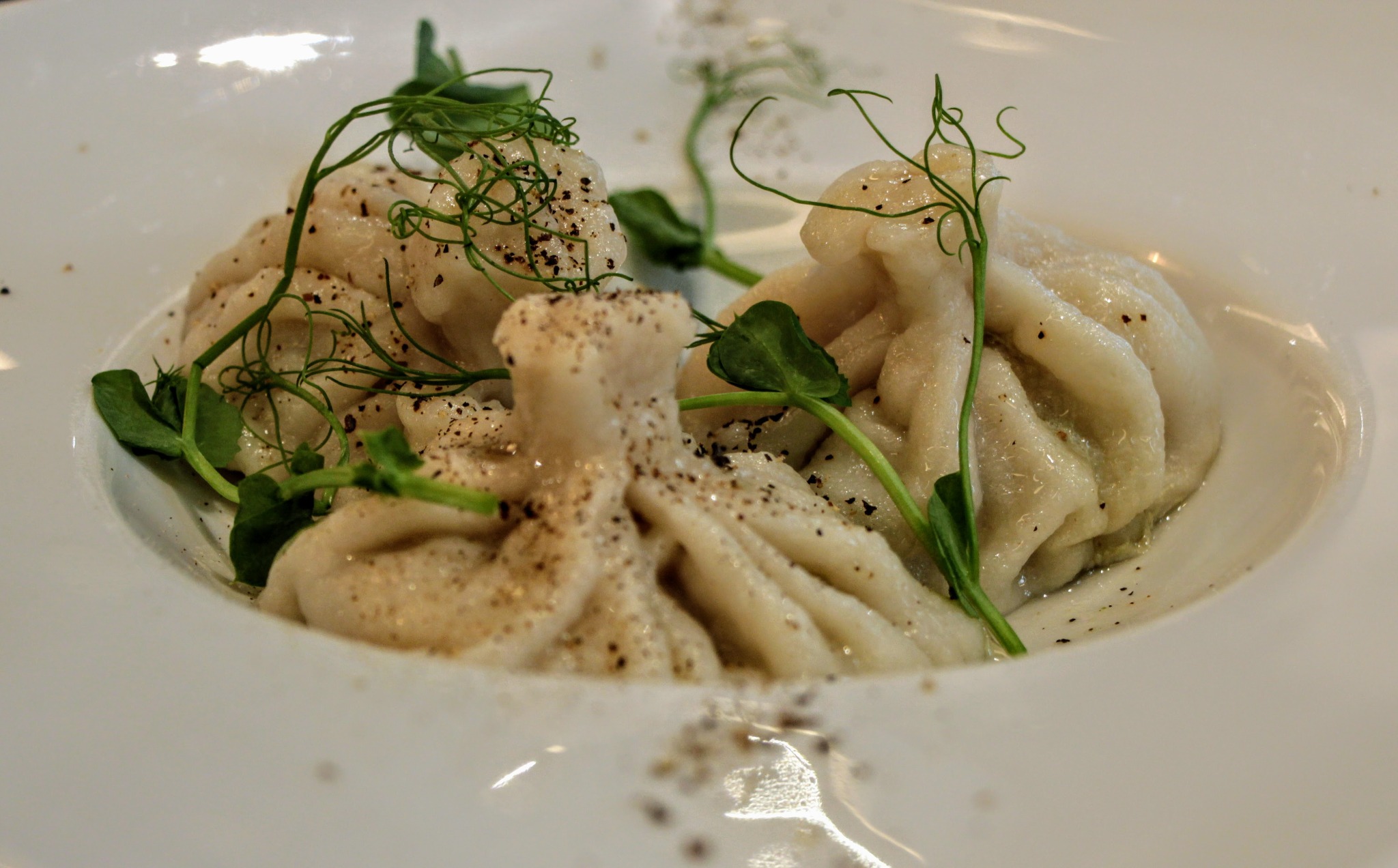 Dwa chinkali posypane pieprzem i udekorowane młodymi pędami groszku, podane na białym talerzu.