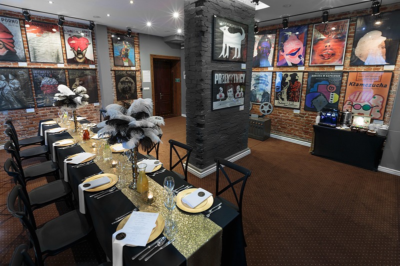 Uroczysty stół z czarnym obrusem, złotym bieżnikiem i piórami, ustawiony w sali z ceglanymi ścianami ozdobionymi plakatami filmowymi.