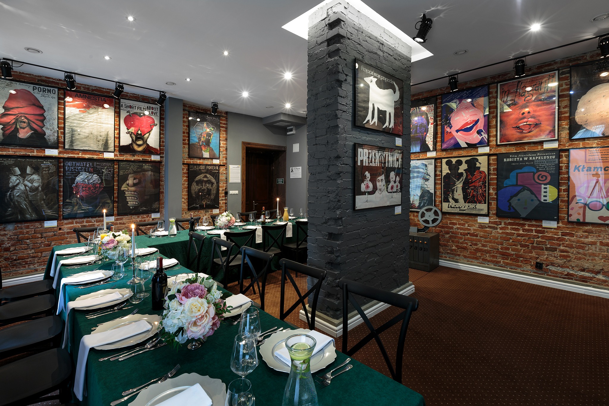 Uroczysta sala z nakrytymi stołami, zielone obrusy, elegancka zastawa, kwiatowe dekoracje, ściany ozdobione plakatami filmowymi, ceglany filar.