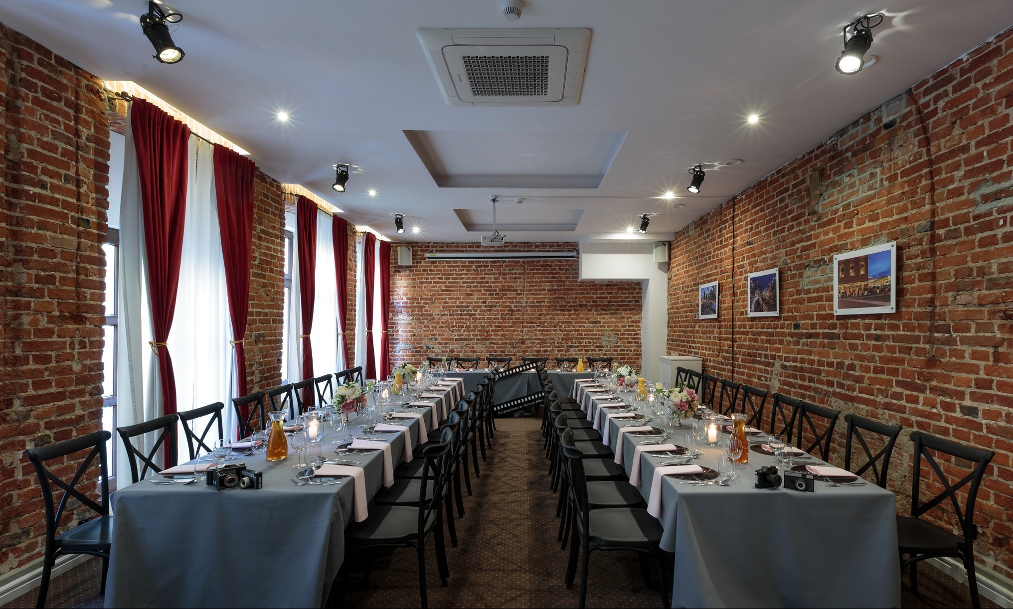 Uroczysta sala z długim stołem nakrytym szarym obrusem, czarne krzesła, dekoracje kwiatowe, ceglane ściany, oświetlenie punktowe i wiszące zdjęcia.