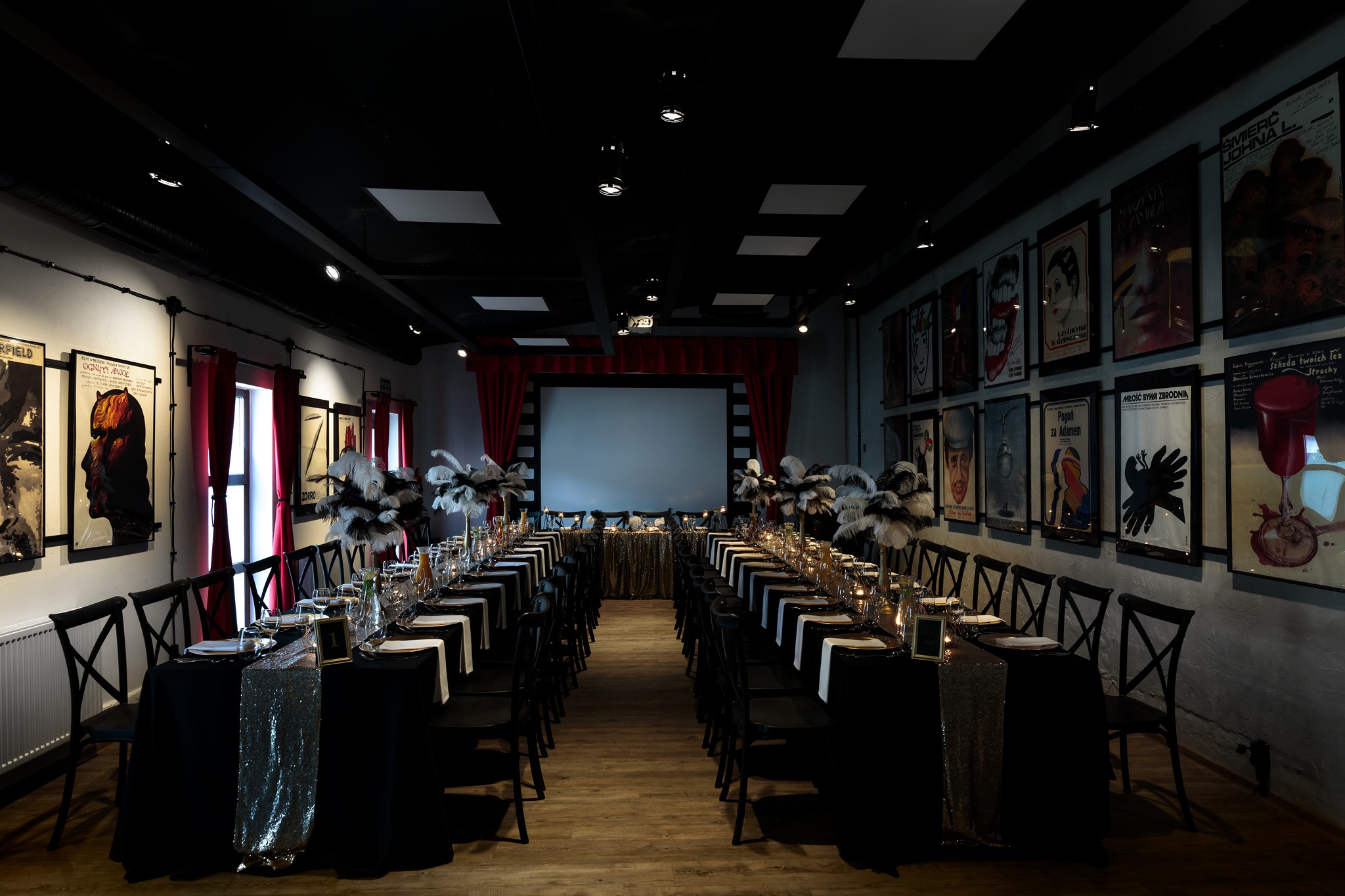Uroczysta sala w stylu retro z długim stołem bankietowym, czarnymi obrusami z błyszczącymi bieżnikami, nakryciami, świecami i dekoracjami z piór, tłem z ekranem i ścianami ozdobionymi plakatami...