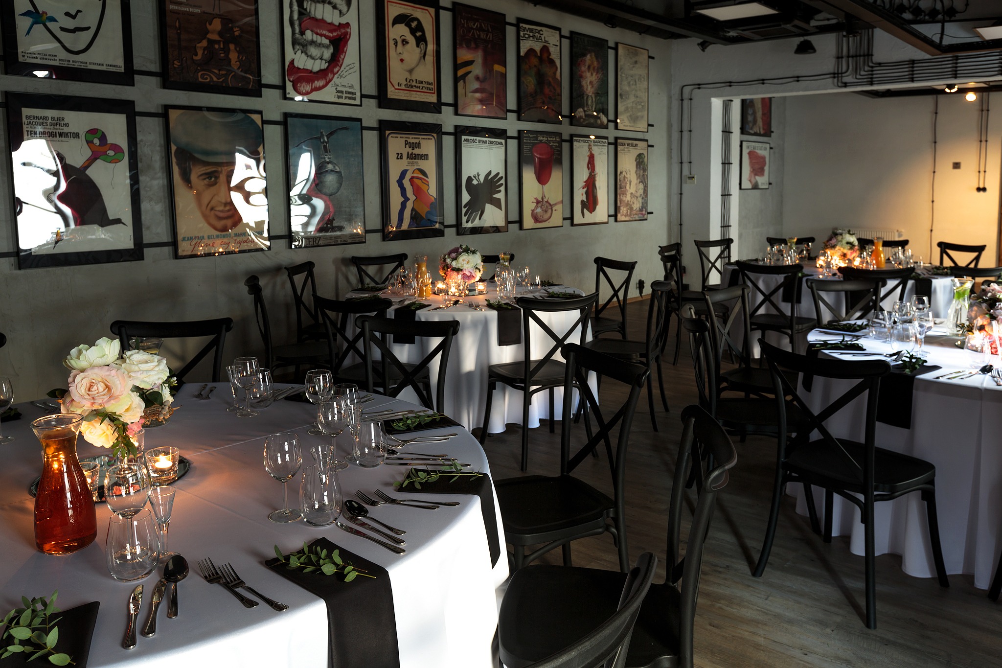 Uroczysta sala z okrągłymi stołami nakrytymi białymi obrusami z czarnymi bieżnikami, czarnymi krzesłami typu crossback, kwiatowymi dekoracjami i świecami, ściany zdobią plakaty filmowe.