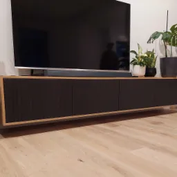 Nowoczesna szafka RTV z czarnymi, ryflowanymi frontami i drewnianą ramą, zawieszona na ścianie, otoczona roślinami doniczkowymi i sprzętem audio, na tle jasnej drewnianej podłogi.