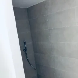 Nowoczesna kabina prysznicowa z szarymi, prostokątnymi płytkami na ścianach i kwadratową deszczownicą w suficie, widok z dołu, z fragmentem białej ściany po lewej stronie.