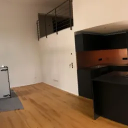 Przestronne wnętrze po remoncie: jasne ściany, drewniana podłoga, nowoczesna czarna kuchnia z miedzianym panelem, okno z czarną ramą i antresola z metalową barierką.