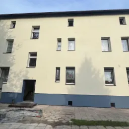 Trzykondygnacyjny budynek po renowacji elewacji z elementami dekoracyjnymi w kolorze brązowym i niebieskim, widoczne okna z firankami i częściowo otwarty garaż z narzędziami.