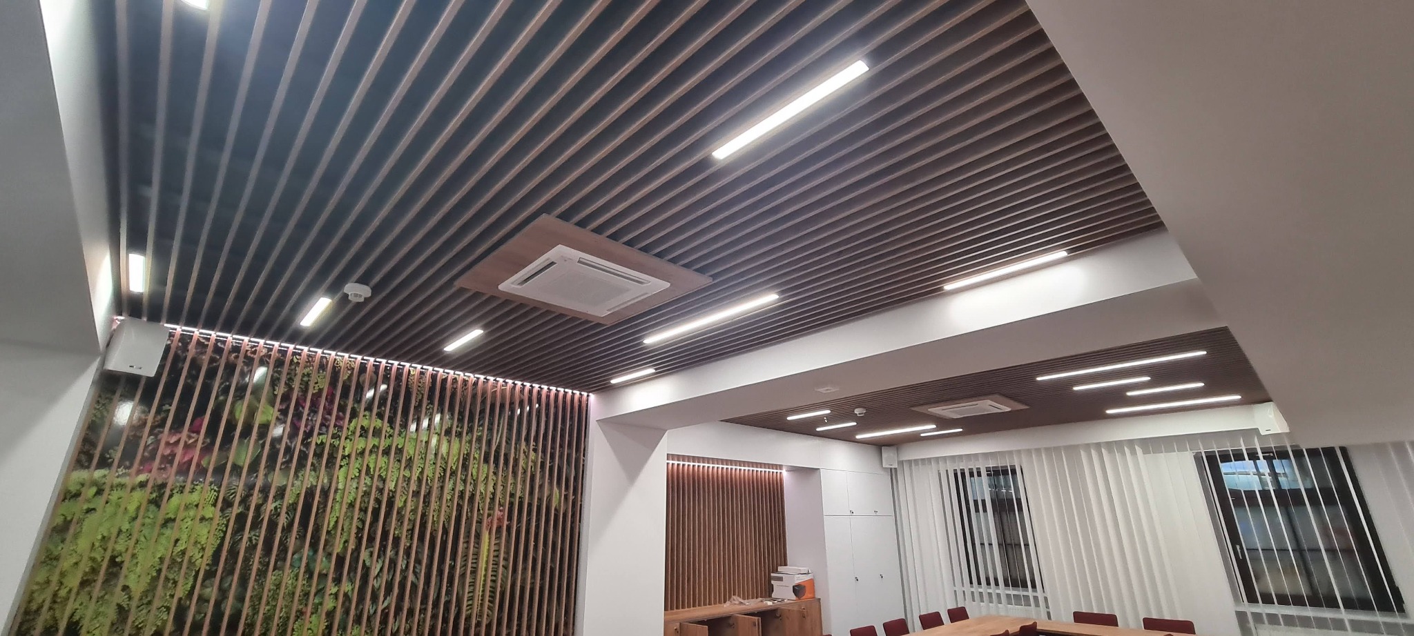 Nowoczesna salka konferencyjna z drewnianym sufitem lamelowym, oświetleniem LED liniowym, klimatyzacją kasetonową i ścianą z zielonym panelem dekoracyjnym.