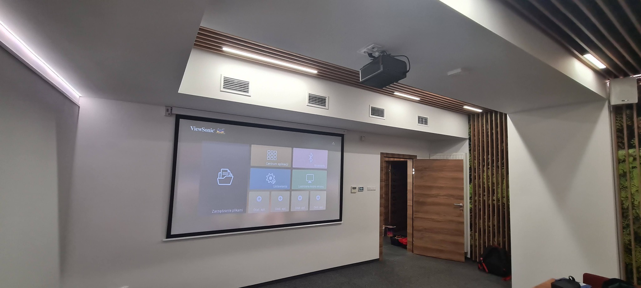 Nowoczesna salka konferencyjna z projektorem sufitowym, ekranem projekcyjnym ViewSonic wyświetlającym menu, oświetleniem LED w suficie oraz dekoracyjną ścianą z drewnianymi listwami.